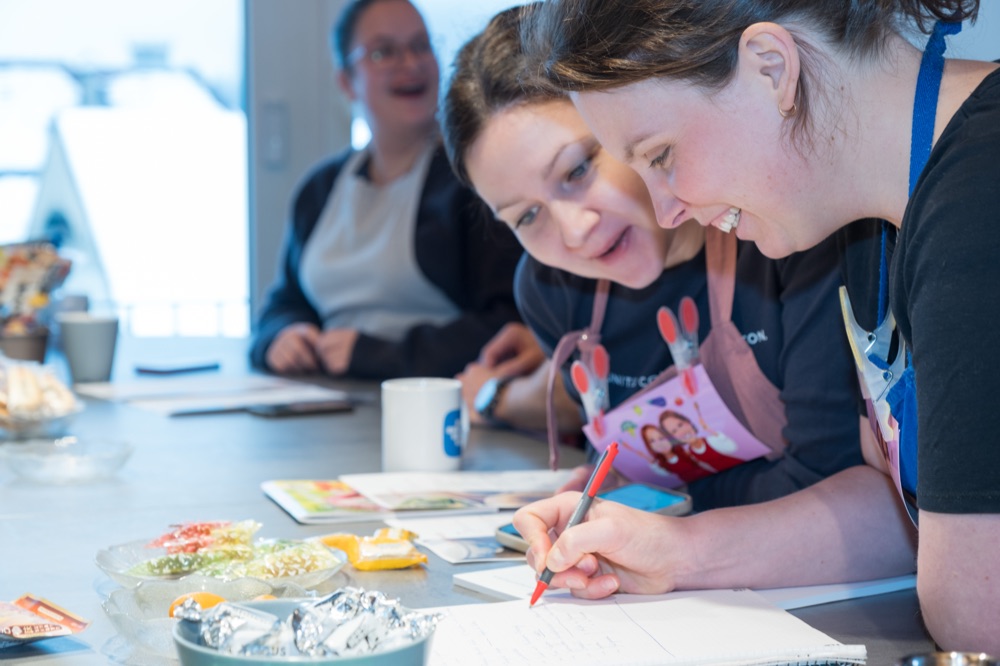 Two workshop participants in aprons laughing while sketching notes together at the kitchen table