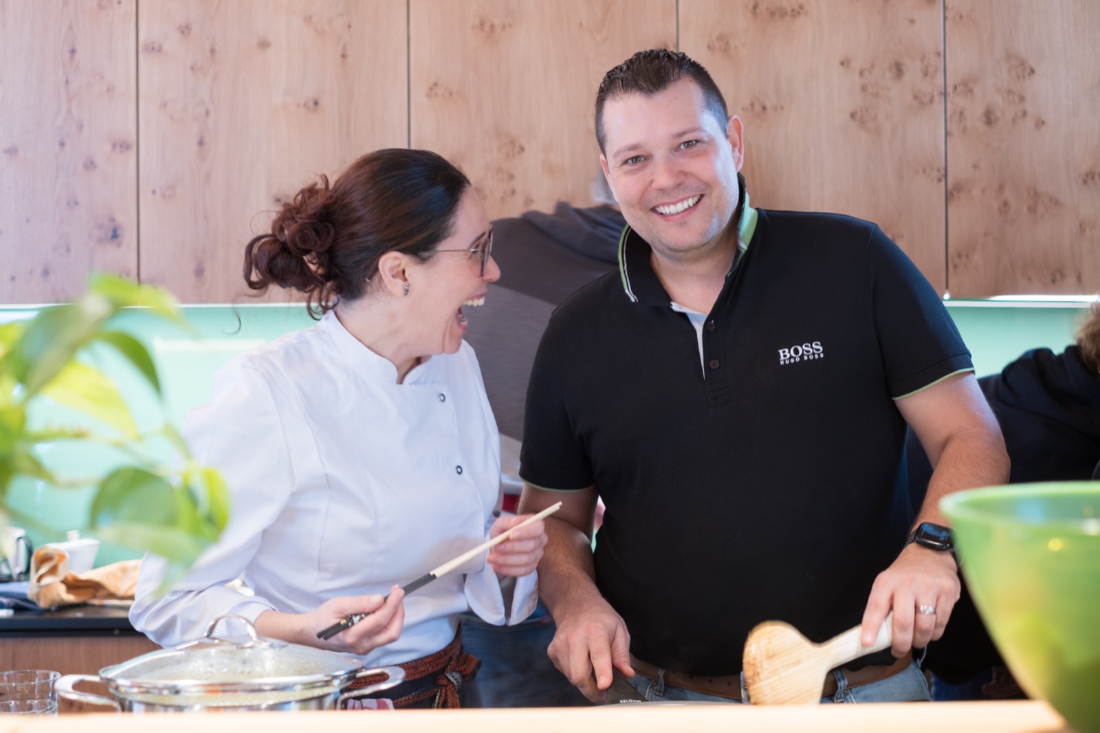 Ophelia in her chef's jacket laughing with a workshop participant at the kitchen counter, wooden spoon and steaming pot at hand