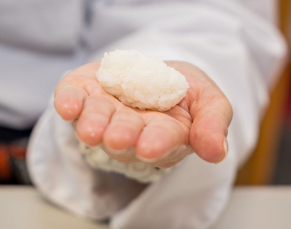 Open hand in a chef's jacket sleeve presenting a freshly formed ball of sushi rice